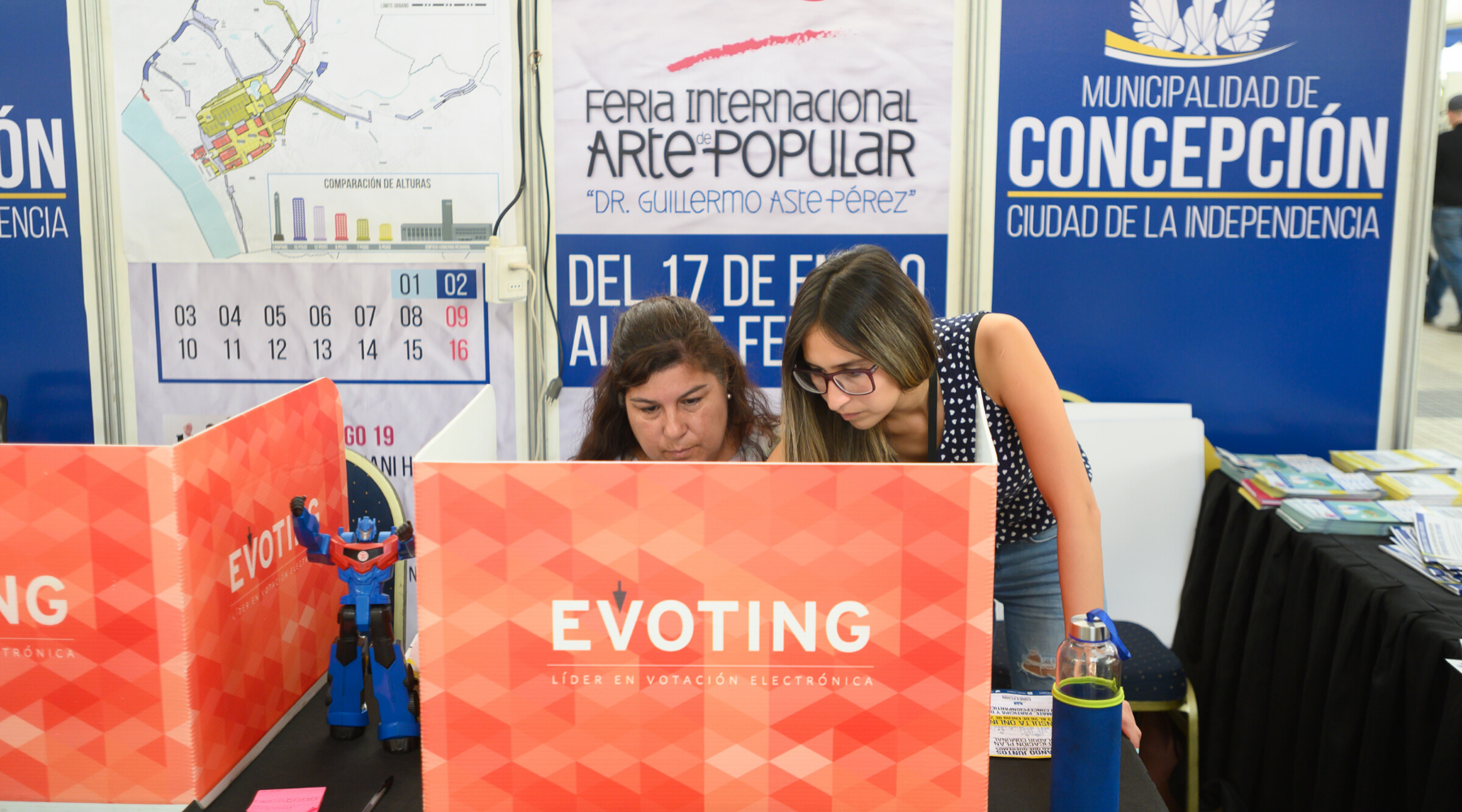 Mujer votando electrónicamente siendo asistida por otra mujer en la Consulta al Plan Regulador de Concepción, con logo de EVoting en la esquina